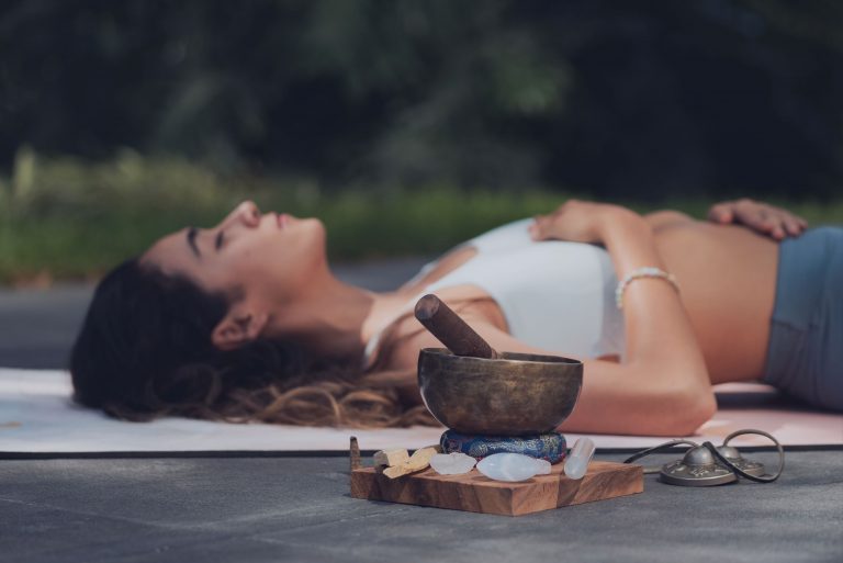 Mujer recostada al aire libre con las manos sobre el abdomen, junto a un cuenco tibetano, cristales y campanas tibetanas, en una sesión de meditación y sanación energética; imagen tomada para Caminar Creciendo, Sevilla.