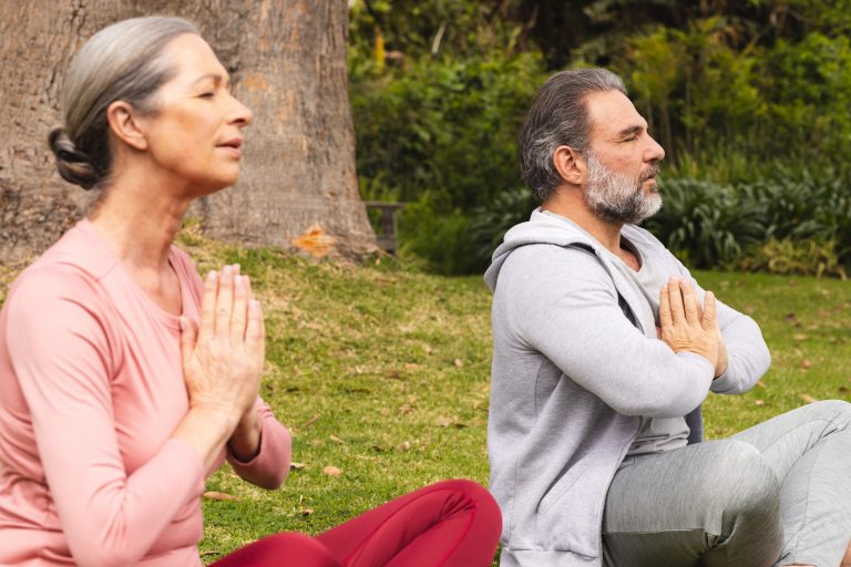 Pareja de adultos mayores practicando meditación juntos en posición de oración sobre césped, rodeados de naturaleza, en una actividad de bienestar guiada por Caminar Creciendo en Sevilla.