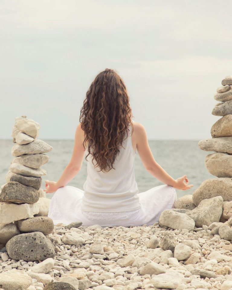 Mujer sentada en postura de meditación sobre piedras blancas frente al mar, entre dos columnas de rocas apiladas; escena serena que transmite equilibrio y conexión con la naturaleza, tomada para Caminar Creciendo, Sevilla.
