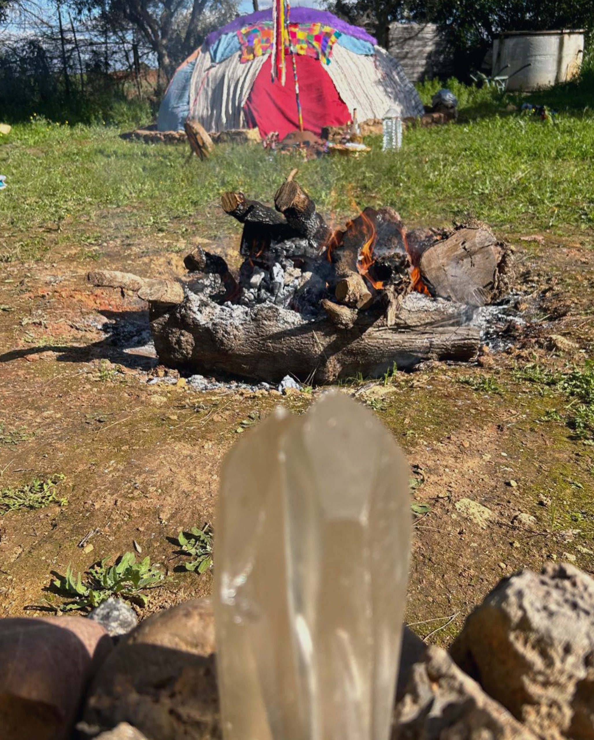 Fuego encendido con leña natural frente a un cristal y una estructura de temazcal decorada con telas coloridas en un espacio verde; imagen representativa de ceremonias ancestrales y conexión espiritual en Caminar Creciendo, Sevilla.