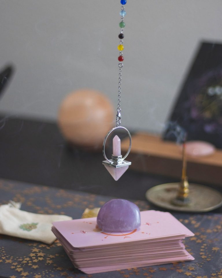 Péndulo con cuentas de colores suspendido sobre un conjunto de cartas energéticas decoradas, junto a una piedra amatista y elementos esotéricos; escena simbólica de prácticas de conexión espiritual en Caminar Creciendo, Sevilla.