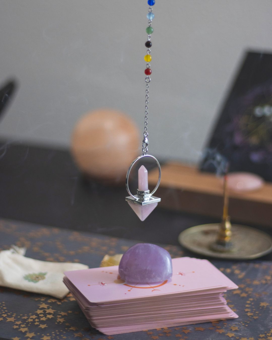 Péndulo con cuentas de colores suspendido sobre un conjunto de cartas energéticas decoradas, junto a una piedra amatista y elementos esotéricos; escena simbólica de prácticas de conexión espiritual en Caminar Creciendo, Sevilla.