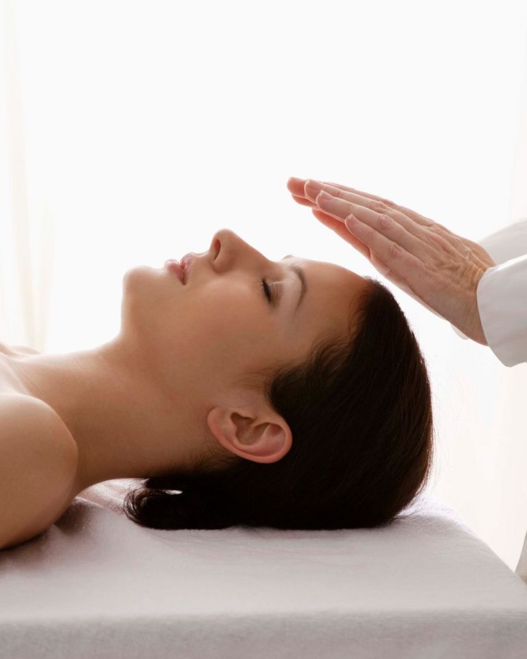 Mujer recostada con los ojos cerrados recibiendo una sesión de reiki, con las manos del terapeuta suspendidas sobre su cabeza en un ambiente sereno; imagen representativa de terapias energéticas en Caminar Creciendo, Sevilla.