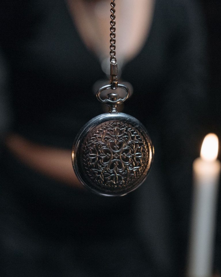 Primer plano de un reloj de bolsillo con grabados colgando de una cadena, utilizado como péndulo en un ambiente tenue con vela encendida; imagen representativa de técnicas de relajación o hipnosis para Caminar Creciendo en Sevilla.